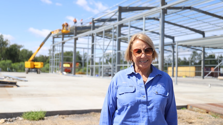 Jenny Hayes stands in the forefront while builders work on shed construction in the background. 