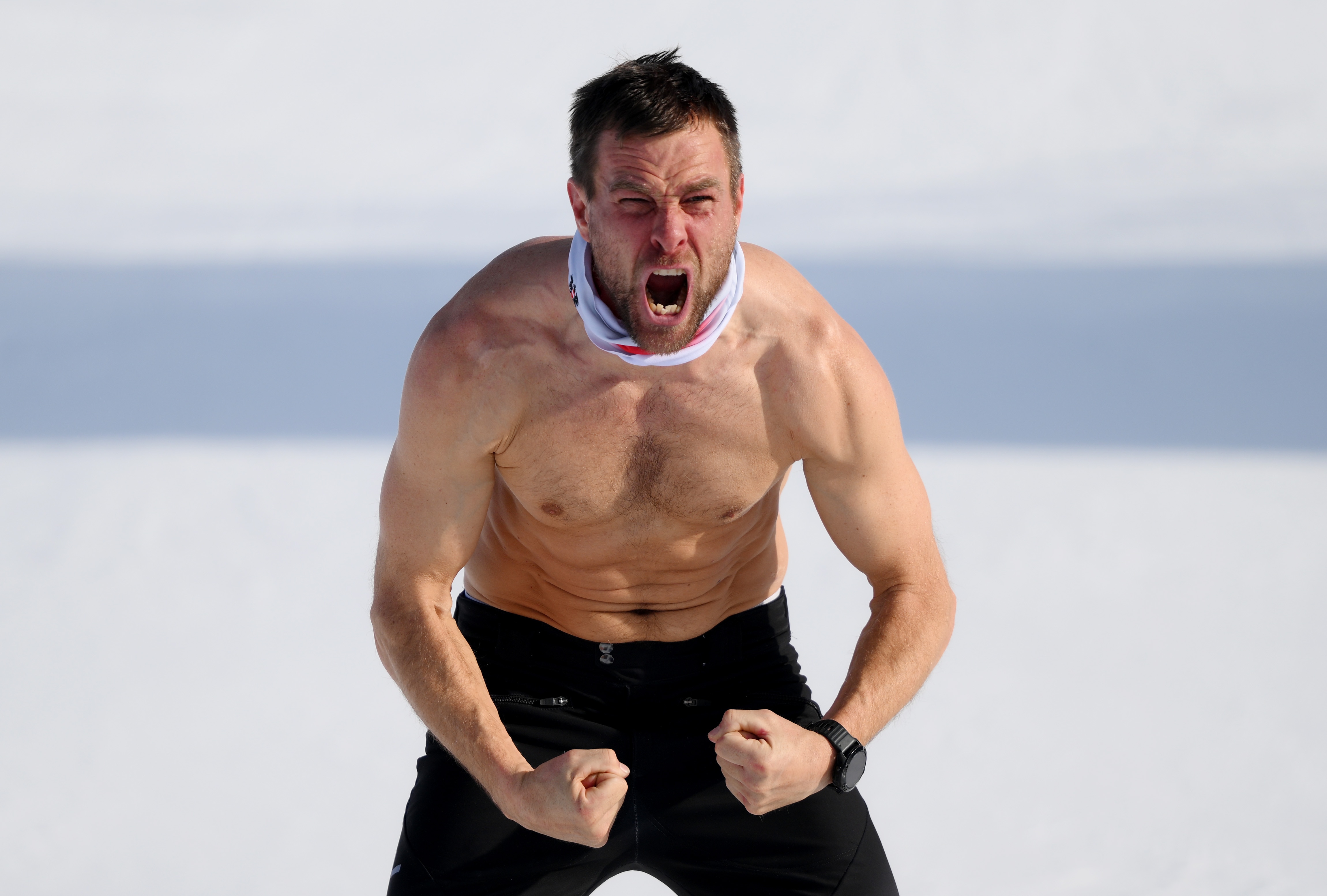 Un hombre en topless con pelo corto y negro flexiona sus músculos en la nieve y grita