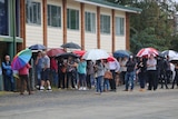 A queue of people stand in the rain with umbrellas.