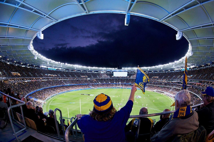 A fisheye shot of a full Perth Stadium at night, with a fan waving an Eagles flag in the foreground