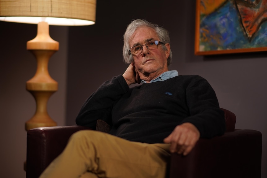 A man sits on a lounge chair with a large wooden lamp beside him