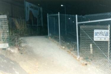 A rough gravel pathway with cyclone fencing, pictured at night.
