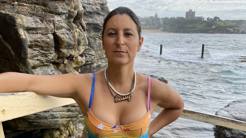 A trans woman wearing a rainbow dress standing in front of ocean baths, with a rock wall on the left.