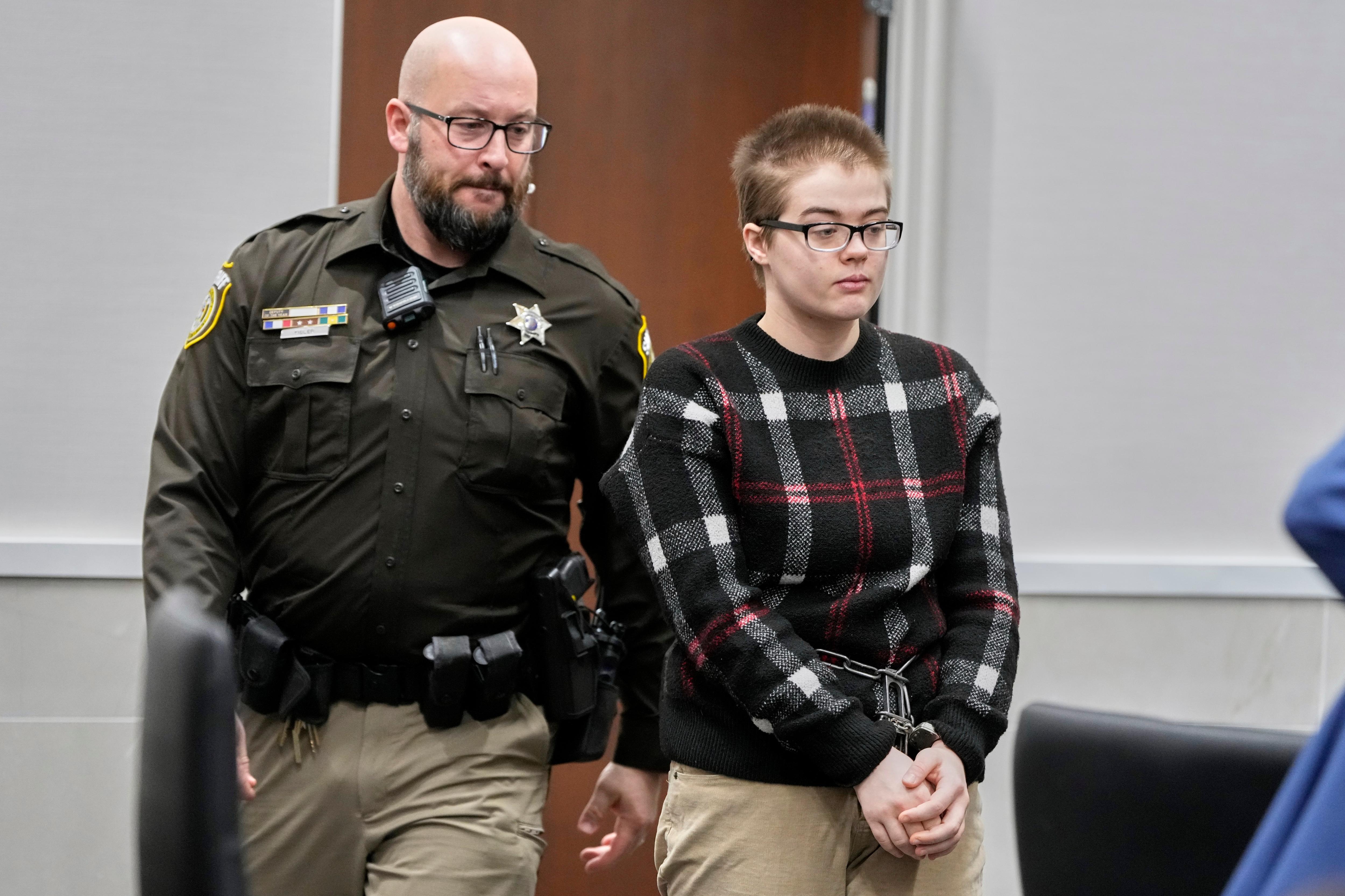 woman is escorted to a seat inside a courtroom by a police man