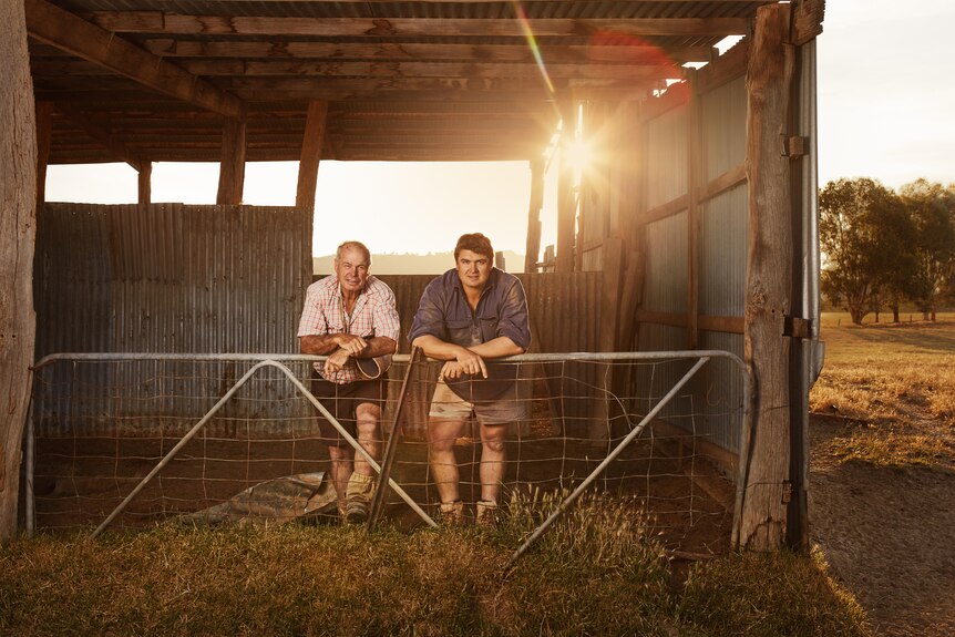 Two men leaning on a barn fence holding hats 