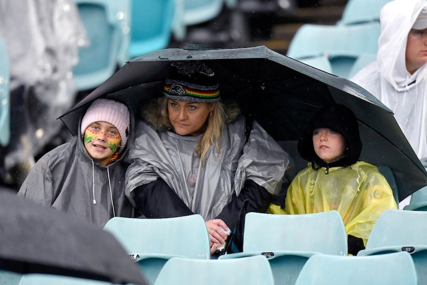 A woman and two children wearing ponchos sit in a stadium under a large umbrella.
