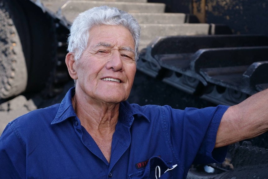 An elderly man smiles at the camera, standing in front of the wheel of a mining truck
