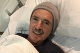 A man lies in a hospital bed with his blanket up to his neck and he is wearing a beanie.