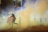 A woman carrying a placard runs through a cloud of yellowish tear gas.