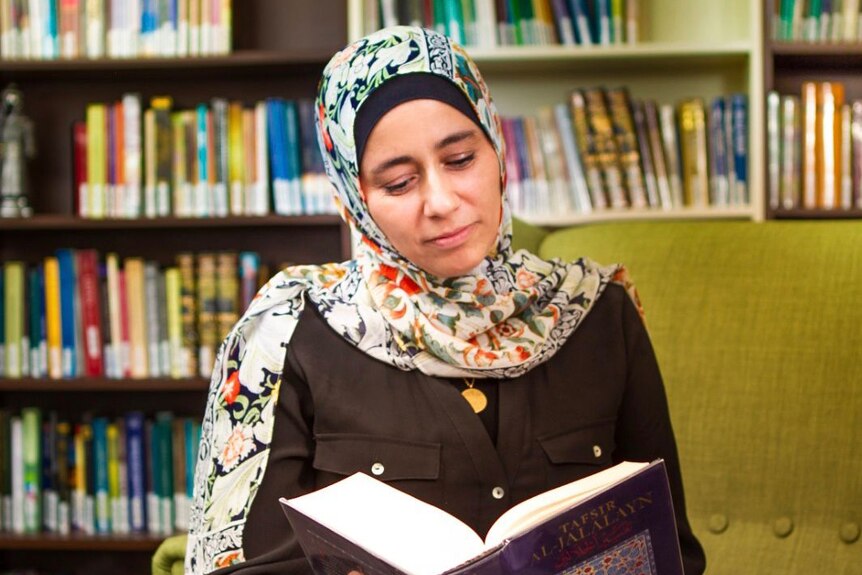 Qur'anic researcher Mouna Elmir wearing floral hijab and reading a book, with bookcases behind her.