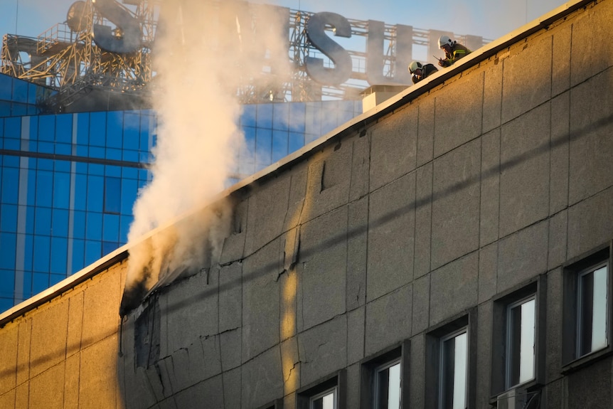 两名消防员站在一栋瓦片碎片和浓烟滚滚的建筑物的屋顶上。