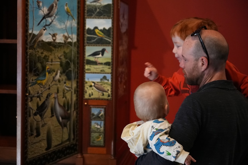 A man holds up two young children to have a look at a painting of birds.