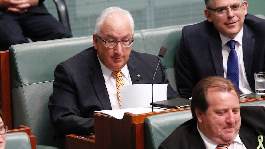 Danby is wearing a yellow tie, holding a piece of paper, looking towards the government benches.