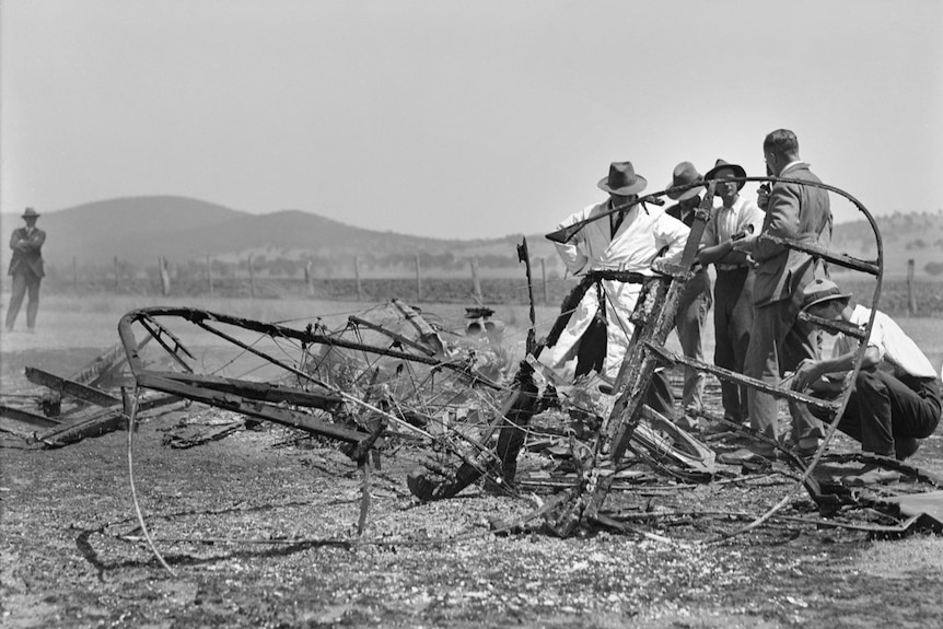 Black and white photo of a plane crash. The plane has been reduced to rubble in a field. Investigators are inspecting the wreck.
