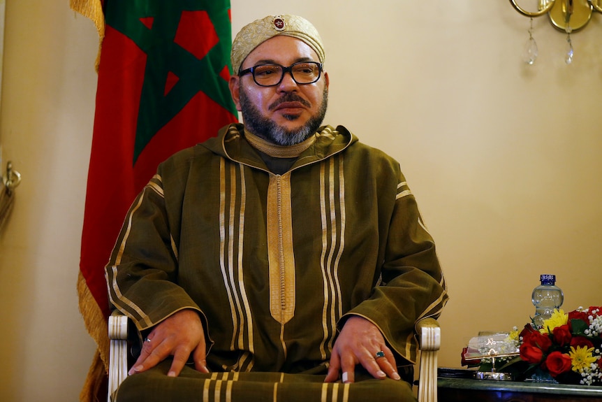 King Mohammed VI of Morocco sits on a chair wearing a green jumper and looking into the distance.