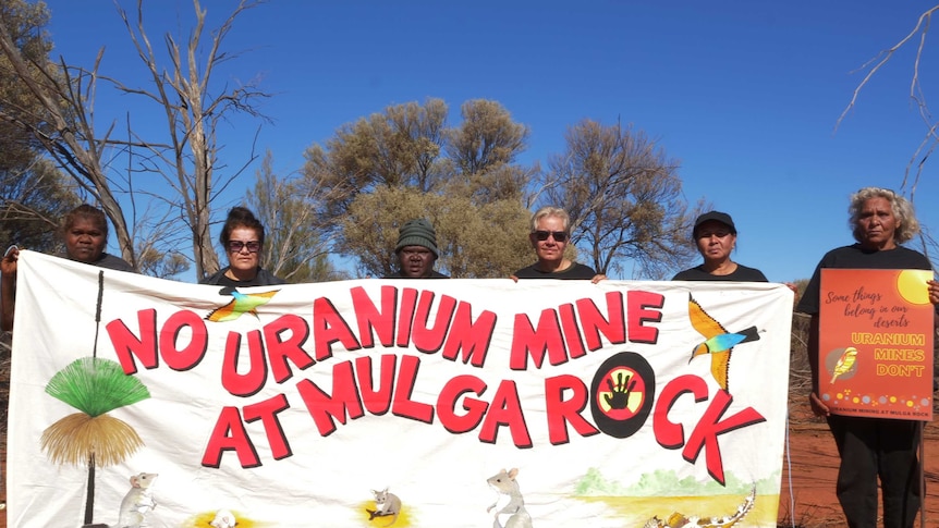 6 Aboriginal women stand on country holding a banner which says 'No uranium mining at Mulga Rock'. One holds a poster