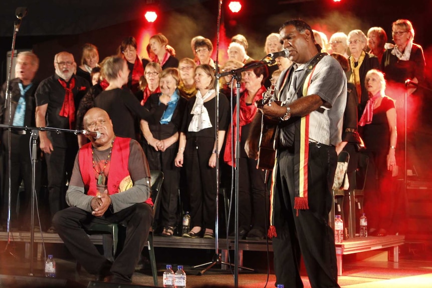 Two Indigenous men perform on stage, one holding a guitar, a choir stands behind.