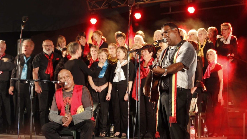 Two Indigenous men perform on stage, one holding a guitar, a choir stands behind.