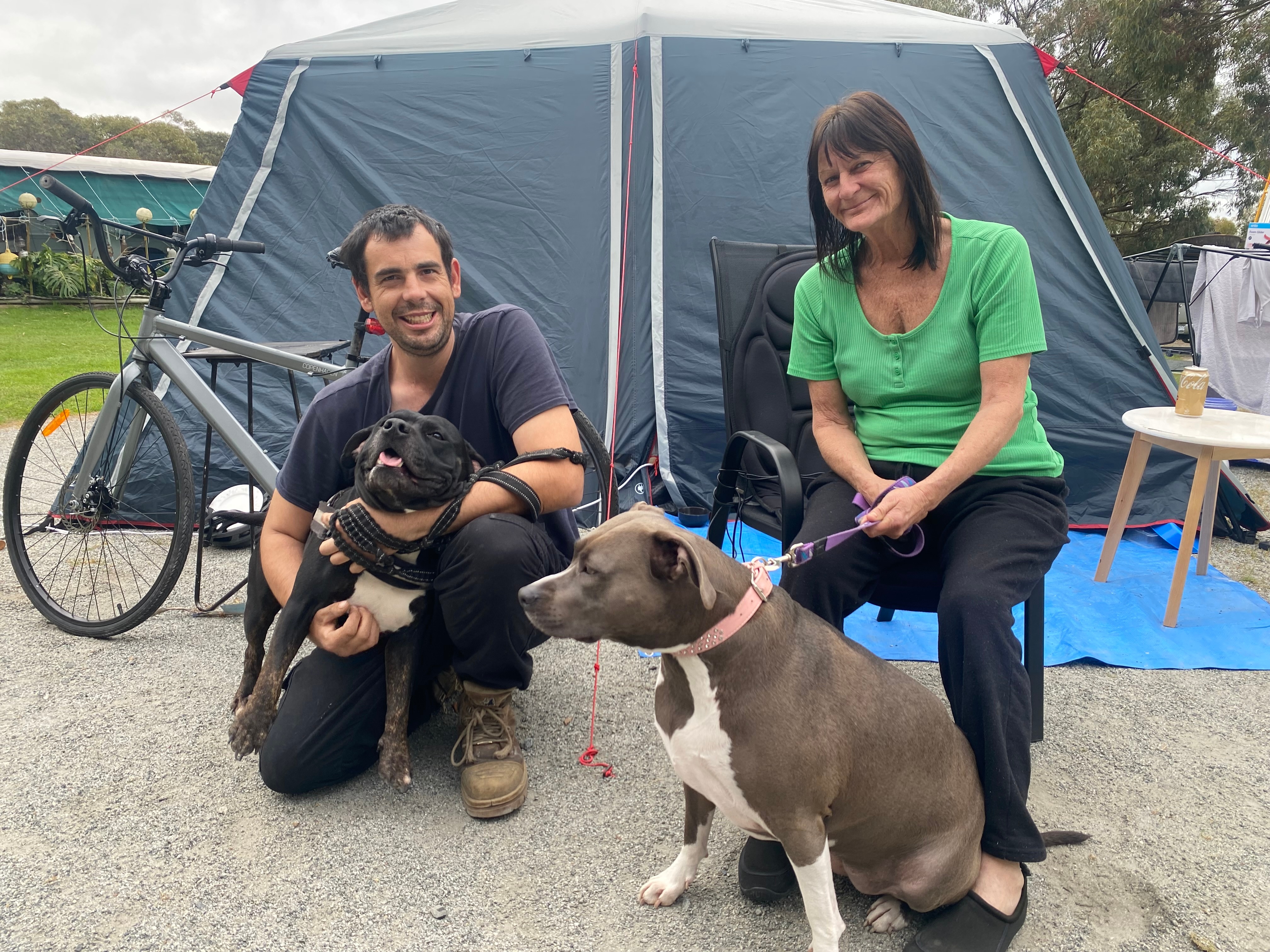 Man and woman holding dogs on leads in front of a tent and bike