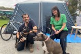A dark-haired man in his 30s and his mother, about 60, sit in front of a tent with their two dogs.