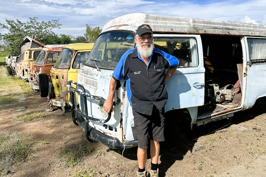 A man with long grey beard, black and blue mechanic's shirt, shorts, stands in front of old Volkswagen vehicles.