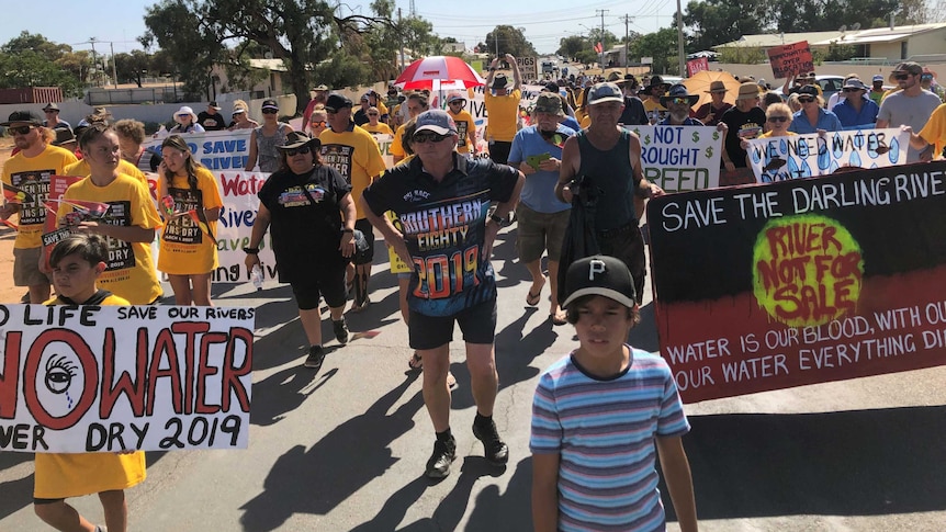 A large group of people walk down a road holding signs reading, 'No life. No water. Save our river'.