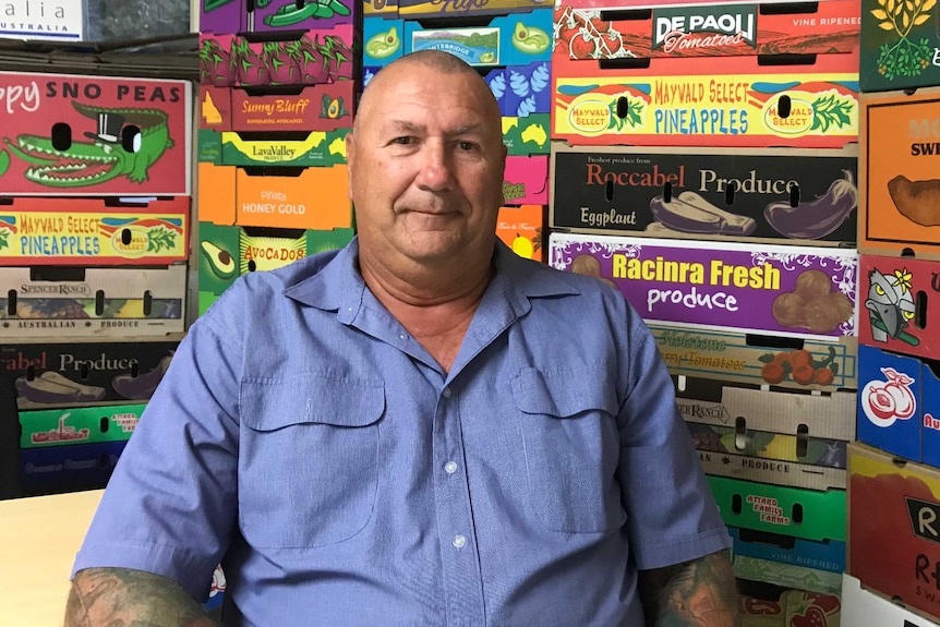 A man in a blue shirt sits in front of stacks of cardboard fruit and vegetable cartons.
