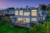 A three story luxury home on a hill with a deck shown at night with lights on