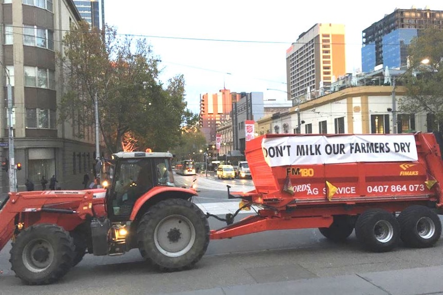 A red tractor with Don't Milk Our Farmers Dry banner on its side blocks a city  road. High rise building in the background.