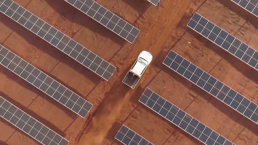 Aerial shot of a white utility dwarfed by rows of solar panels against a red dirt background.