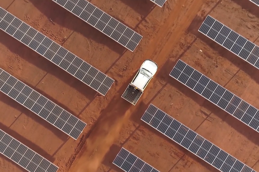 Aerial shot of a white utility dwarfed by rows of solar panels against a red dirt background.