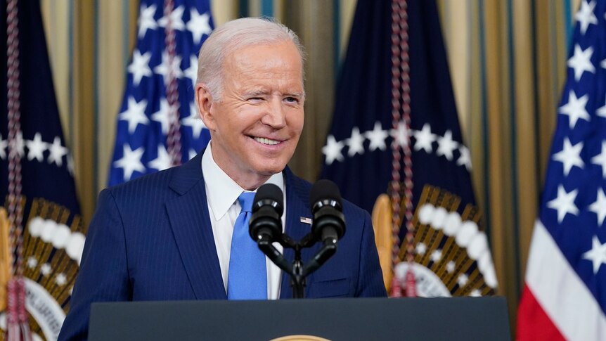 Joe Biden in dark suit at presidential podium smiles and winks slightly