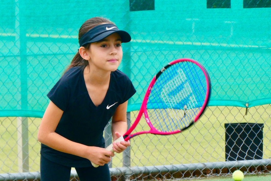 A young girl stands ready to receive a ball with a pink tennis racket.