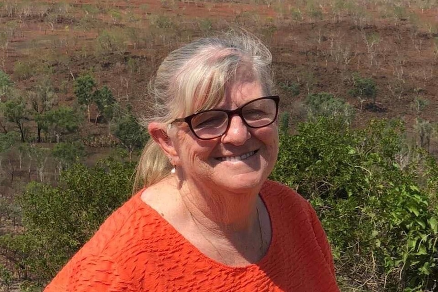 An older woman wearing glasses and an orange top smiles for the camera.