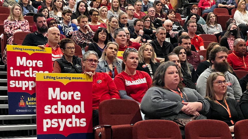 A group of people, some holding placards, sit in a lecture theatre.