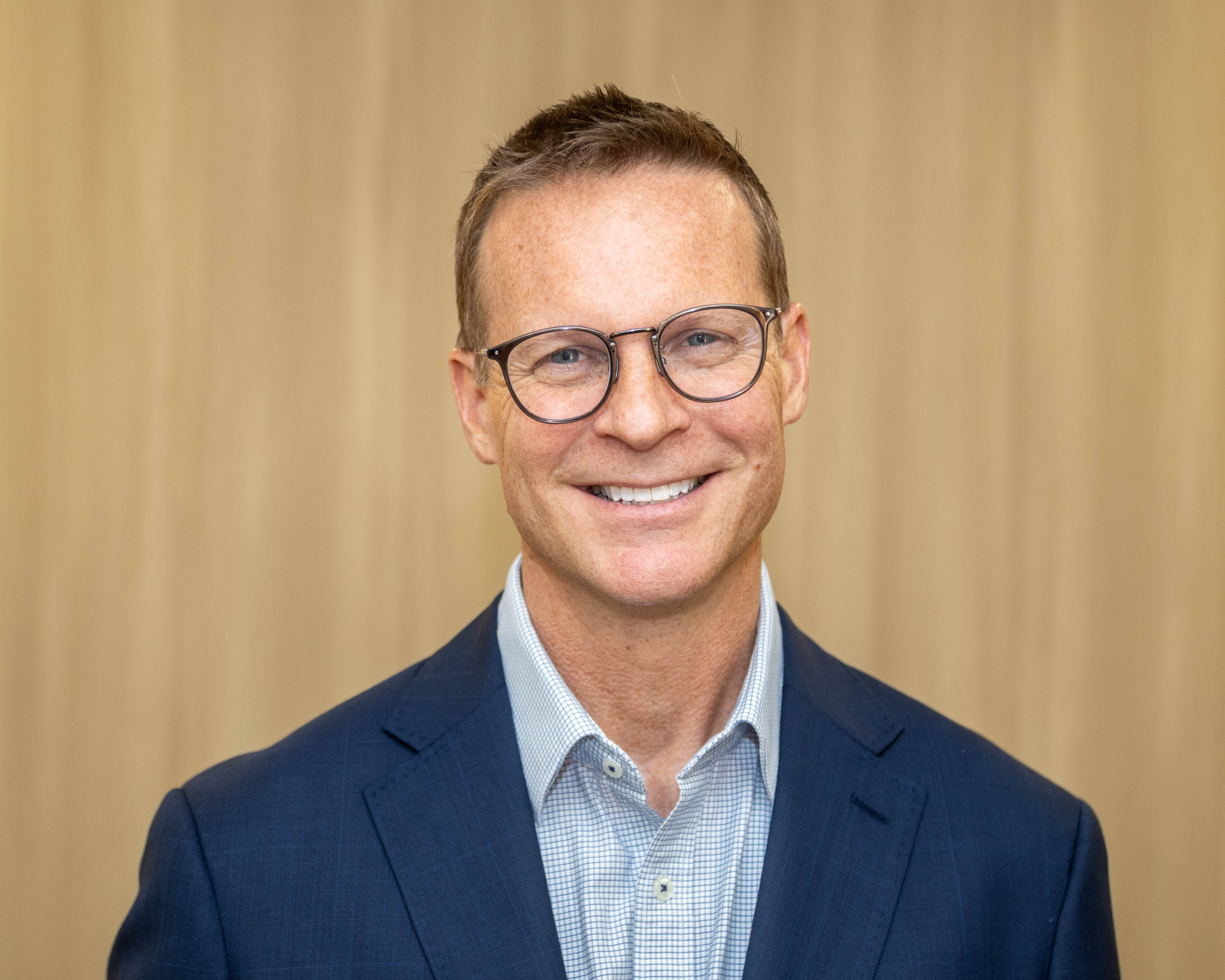 A man with sandy hair wearing glasses, a navy jacket and blue collared shirt smiles at the camera with a wood panel backdrop.