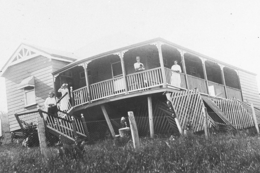 A house with fence and columns slanting with people on the verandah