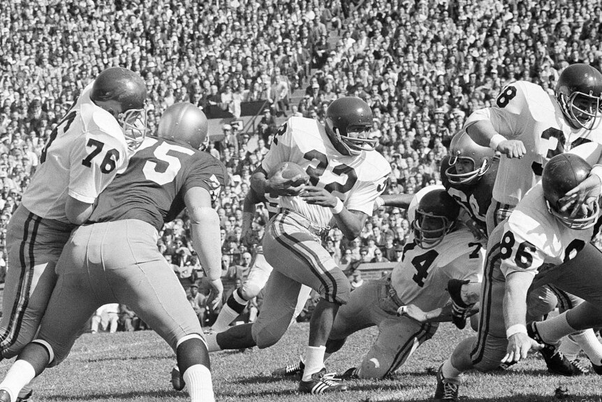 A black and white image of men playing American football in a stadium in front of a big crowd, OJ Simpson runs holding the ball