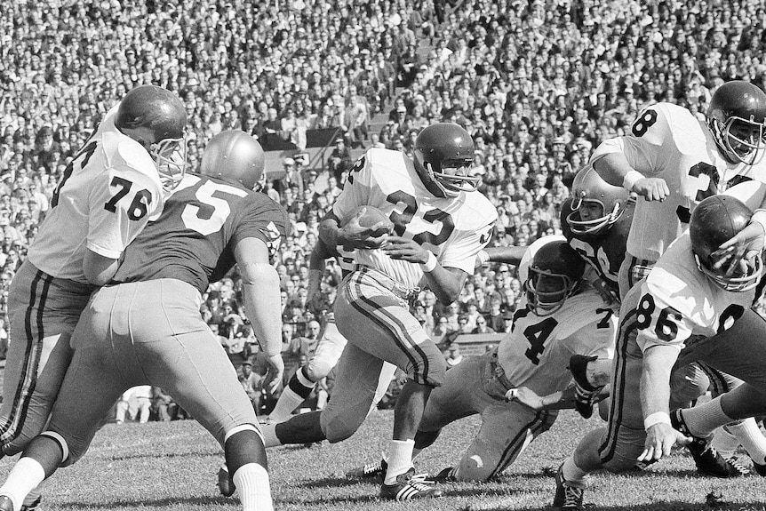 A black and white image of men playing American football in a stadium in front of a big crowd, OJ Simpson runs holding the ball