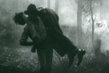 Black and white picture of a man carrying another man over his back in the bush.