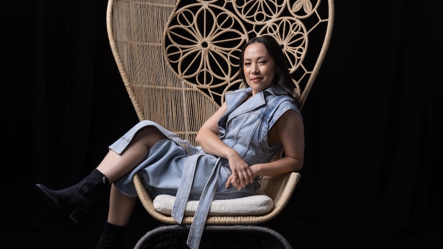 Asian Australian woman with dark hair wears an assymmetric denim dress and sits in an elaborately woven cane arm chair.