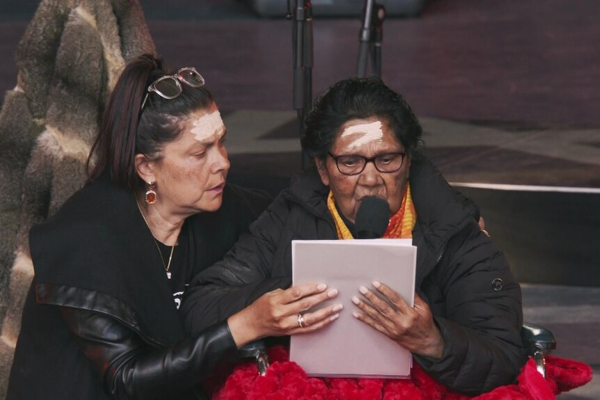Two women sit close, with one reading into a microphone from sheets of paper.