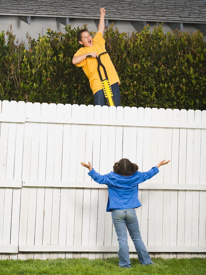A boy jumps over a fence raising his arm while a girl stands below 