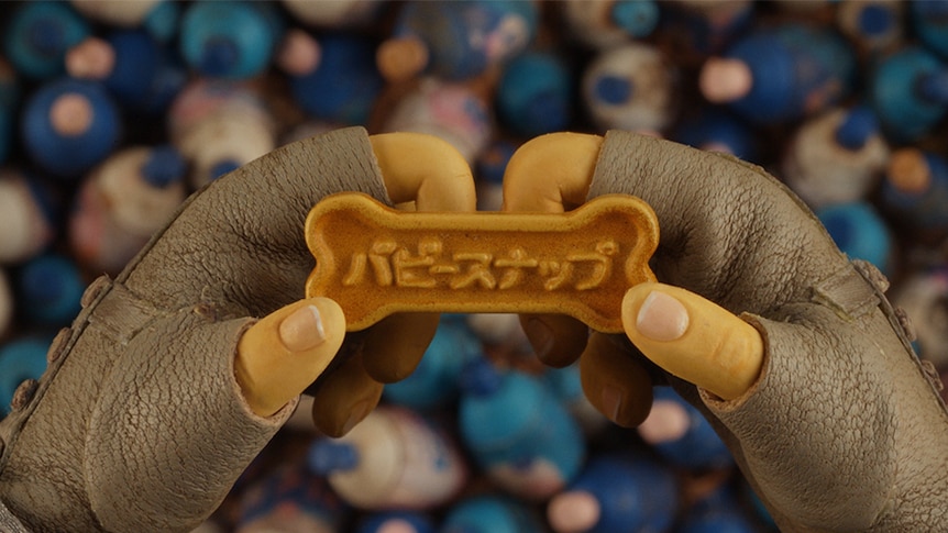 Colour close-up still of puppy snaps dog biscuit held in a puppet's leather glove wearing hand in stop-motion film Isle of Dogs.