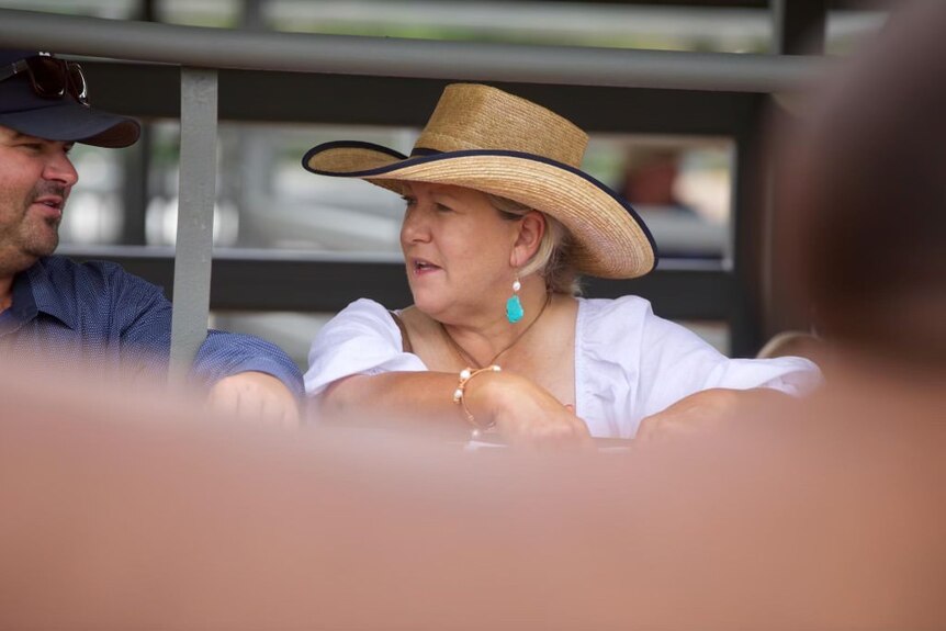 A woman wearing a large straw hat talks to a man. She's dressed in a nice white top and colourful jewelry.