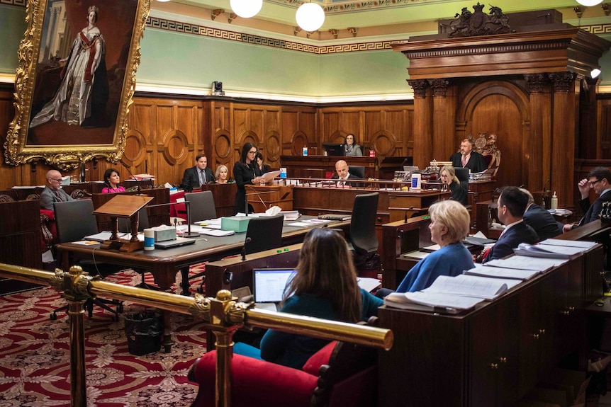 A woman stands at a lectern speaking surrounded by other people sitting.