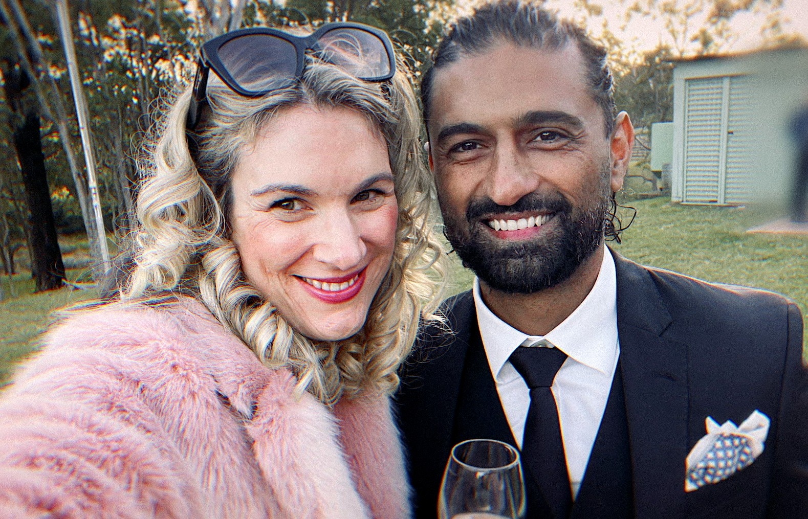 Woman in pink fluffy jacket and sunglasses on head smiles in selfie next to man wearing suit and tie.