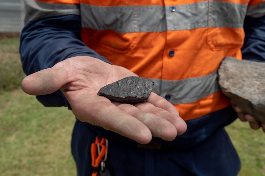 A man holds out his hand with a small lump of coal in it. He is wearing hi-vis workwear.