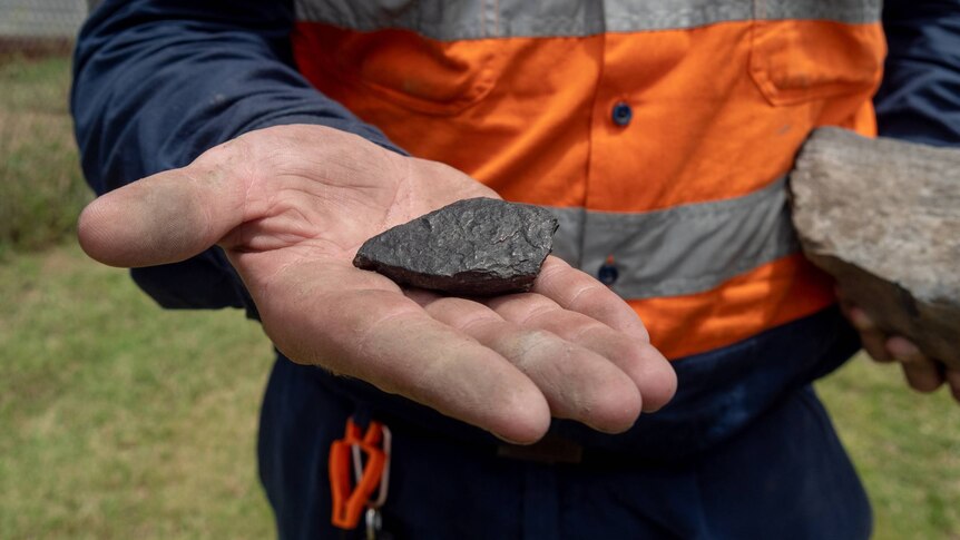 A man holds out his hand with a small lump of coal in it. He is wearing hi-vis workwear.