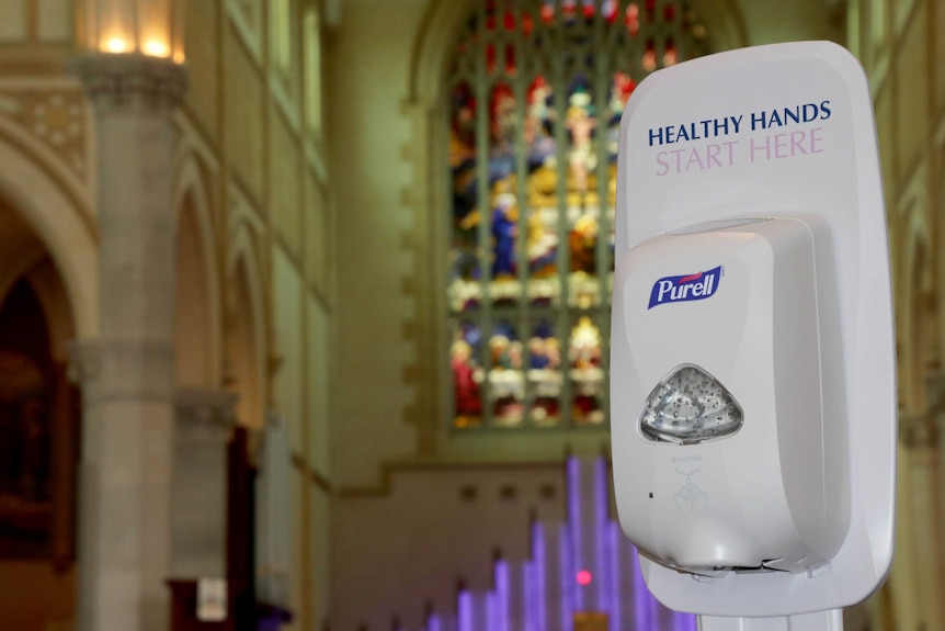 A hand sanitiser in front of the main hall, stained glass window visible in background.